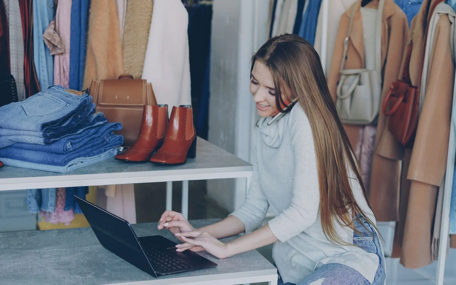 Boutique owner updating her online store on a laptop among racks of clothing